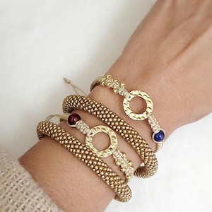May include: Gold-toned bracelets with textured, beaded bands and circular centerpieces. Some bracelets feature small, colorful beads. The bracelets are stacked on a wrist, showcasing their design and style.