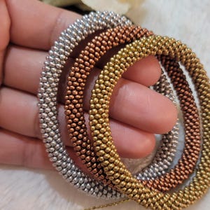 Può includere: Tre braccialetti di perline nei toni argento, rame e oro. I braccialetti sono rotondi e sembrano essere fatti di piccole perline metalliche. È visibile anche una catena d'oro. I braccialetti sono probabilmente gioielli.