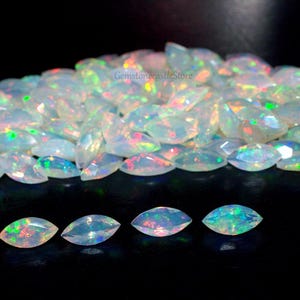 May include: A collection of marquise-cut opals, displaying a milky white base with iridescent flashes of color. The gemstones are scattered across a black surface, with a few stones isolated at the bottom.
