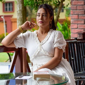 Puede incluir: Una mujer con un vestido blanco con detalles de encaje se sienta en una mesa de madera con tapa de vidrio. Ella sostiene un libro titulado "The Power of Now" y un bolígrafo. La mesa está rodeada de sillas de mimbre.