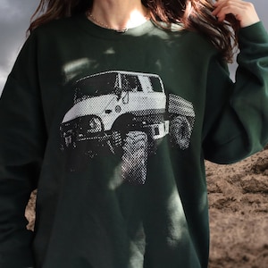 May include: A dark green sweatshirt featuring a white graphic print of a vintage truck. The truck is shown in a side view and the graphic is in a pixelated style.