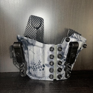 Könnte beinhalten: Ein schwarzes und graues, gemustertes Holster mit zwei schwarzen Clips und einer Rückwand aus Kohlefaser. Das Holster hat eine Reihe schwarzer Schrauben an den Seiten.