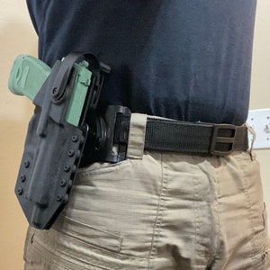 May include: A black holster with a green handgun attached to a black belt. The holster has multiple adjustable retention screws.