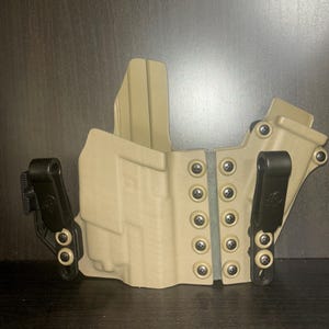 Könnte beinhalten: Ein braunes Lederholster mit schwarzen Clips und verstellbaren Retentionsschrauben. Das Holster ist für das Tragen einer Schusswaffe konzipiert.