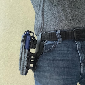 Puede incluir: Una funda de arma negra y azul con un diseño texturizado. La funda está unida a un cinturón negro y se lleva en el lado derecho de la cadera.