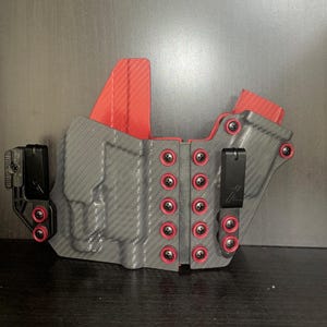 Könnte beinhalten: Ein schwarzes und graues Waffengehäuse mit roten Akzenten. Das Gehäuse hat ein Karbonfaser-Muster und ist aus strapazierfähigem Material gefertigt. Es verfügt über einen Clip zum Befestigen an einem Gürtel und eine Vielzahl von Schrauben zur Anpassung.