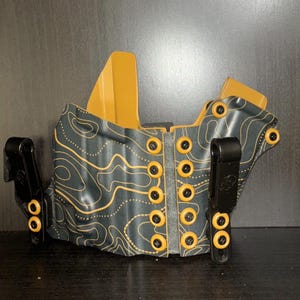 Könnte beinhalten: Ein schwarzes und braunes Lederholster mit topografischem Muster und zwei schwarzen Clips. Das Holster hat insgesamt 12 goldfarbene Druckknöpfe.