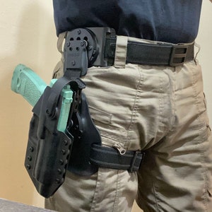 May include: A black holster with a light green handgun attached to a black belt with a black and silver buckle. The holster is secured to the belt with a black strap.