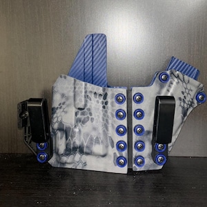 Puede incluir: Una funda de pistola negra y gris con un patrón de camuflaje y detalles azules. La funda tiene un clip en el lateral y un bucle en la parte posterior.