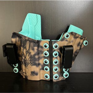 Könnte beinhalten: Ein Holster in Tarnmuster in Schwarz und Braun mit hellblauem Innenfutter und schwarzen Clips. Das Holster hat eine Reihe von türkisfarbenen Knöpfen in der Mitte.