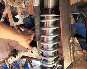 May include: A close-up of a silver shock absorber with a black spring and a black hose attached to it. The shock absorber is mounted to a metal bracket with several holes drilled into it. The bracket is attached to a rusty metal frame.