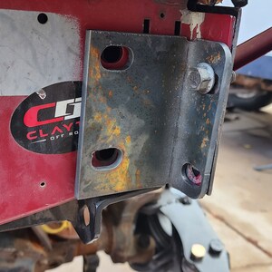 K&ouml;nnte beinhalten: Nahaufnahme einer rostigen Metallhalterung, die an einer roten Metalloberfl&auml;che befestigt ist. Die Halterung hat mehrere L&ouml;cher und ist mit einer Schraube befestigt. Die Oberfl&auml;che hat einen Aufkleber mit dem Text "CLAYTON OFF ROAD" und einen Barcode mit den Nummern "0486482" und "XL592827 3812".