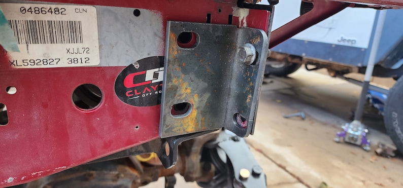 K&ouml;nnte beinhalten: Nahaufnahme einer rostigen Metallhalterung, die an einer roten Metalloberfl&auml;che befestigt ist. Die Halterung hat mehrere L&ouml;cher und ist mit einer Schraube befestigt. Die Oberfl&auml;che hat einen Aufkleber mit dem Text "CLAYTON OFF ROAD" und einen Barcode mit den Nummern "0486482" und "XL592827 3812".