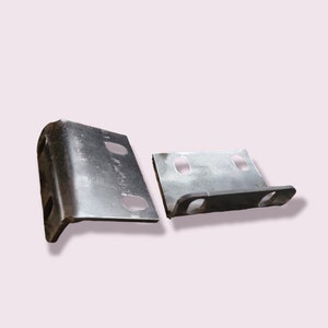 K&ouml;nnte beinhalten: Zwei Metallwinkel mit L&ouml;chern f&uuml;r Schrauben. Die Winkel sind silberfarben und haben eine leicht gebogene Form.