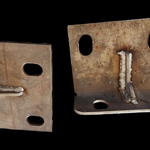 K&ouml;nnte beinhalten: Zwei Metallwinkel mit geschwei&szlig;ten Verbindungen und mehreren L&ouml;chern. Die Winkel sind aus rauh, braunem Metall.