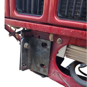 K&ouml;nnte beinhalten: Nahaufnahme der vorderen Sto&szlig;stange eines roten Autos mit einem befestigten Metallhalter. Der Halter hat eine Schraube und einige L&ouml;cher. Die Sto&szlig;stange hat einen schwarzen Kunststoffgrill und ein silbernes Metallteil.