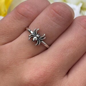 May include: A silver ring featuring a spider design. The ring is worn on a finger, with the spider prominently displayed. The background includes yellow and white flowers, adding a contrasting backdrop to the jewelry.