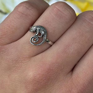 Puede incluir: Un anillo de plata con un diseño detallado de camaleón. El lagarto tiene escamas texturizadas y una cola enroscada. El anillo se lleva en un dedo, sobre un fondo de flores blancas y amarillas. Una joya.