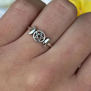 May include: A silver ring featuring a pentagram design in the center, flanked by crescent moon shapes. The ring is worn on a finger, with a background of yellow and white flowers.