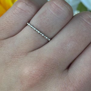 May include: A silver ring with a beaded design is worn on a finger. The ring is thin and delicate, with small, evenly spaced beads creating a textured surface. The hand is set against a backdrop of yellow and white flowers.