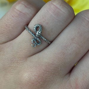 Puede incluir: Un anillo plateado con forma de serpiente enrollada alrededor de un dedo. El anillo tiene una superficie texturizada y la cabeza de la serpiente es visible. El fondo presenta flores amarillas y blancas.