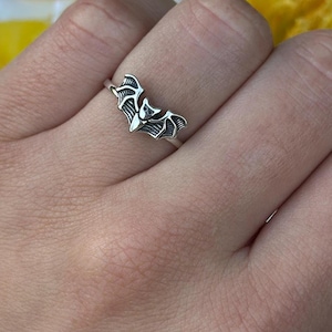 May include: A silver ring featuring a bat design. The bat has outstretched wings and is centered on the ring. The ring is worn on a finger, with a background of yellow and white flowers.