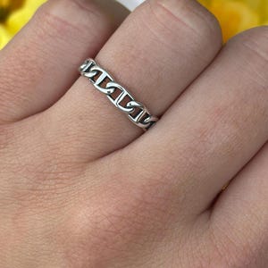 May include: A silver chain link ring is displayed on a hand. The ring has a continuous chain design, with each link connected to the next. The hand is set against a backdrop of yellow and white flowers.