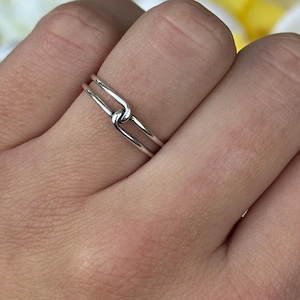 May include: A silver ring with a knot design. The ring is thin and delicate, with a simple yet elegant style. The ring is worn on a finger, showcasing its minimalist aesthetic.