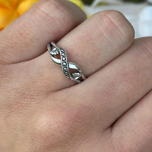 May include: A silver-toned ring with an infinity symbol design. The ring features a twisted band with small, dark-colored stones set along one side. The ring is worn on a finger, with yellow and white flowers in the background.