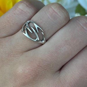 May include: A silver ring with an open, abstract design. The ring features a series of curved lines and shapes, creating a unique and modern look. The ring is shown on a hand with a floral background.