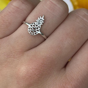 May include: A silver-colored ring featuring a pineapple design. The ring has a detailed pineapple silhouette with a textured body and a leafy crown. The ring is worn on a finger, showcasing the jewelry against a background of yellow flowers.