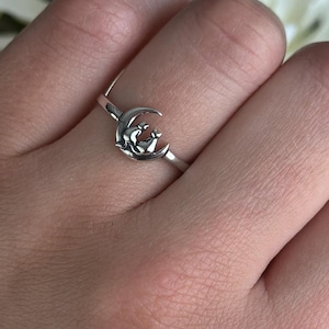 May include: A silver ring featuring a crescent moon design with a small star. The ring is worn on a finger, with a background of yellow and white flowers.