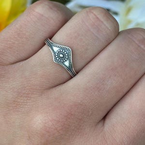 May include: A silver ring with a diamond-shaped design featuring a central floral pattern. The ring is worn on a finger, showcasing its intricate details and craftsmanship. The ring's design is highlighted against a backdrop of yellow and white flowers.