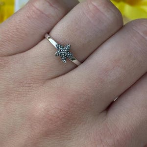 May include: A silver ring featuring a small, textured starfish design. The ring is set against a backdrop of yellow and white flowers. The starfish is approximately 0.25 inches in diameter.