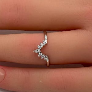 Peut inclure: Une bague en forme de V de couleur argentée ornée d'une série de petites pierres claires taille brillant serties le long de l'anneau en chevron. La bague est portée au doigt, mettant en valeur son design délicat et sa finition métallique.