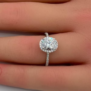 Puede incluir: Un anillo de tono plateado con una gran piedra central de corte ovalado rodeada por un halo de piedras más pequeñas de corte redondo. La banda delgada también está incrustada con diminutas piedras brillantes en su superficie visible.