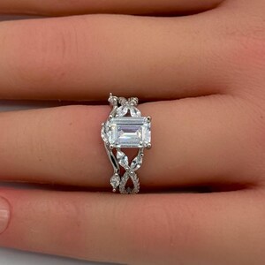 May include: A silver-toned ring featuring a central emerald-cut clear stone flanked by smaller marquise-cut stones arranged in a leaf-like pattern along the band. The metal band has a polished finish and holds multiple clear accent stones.