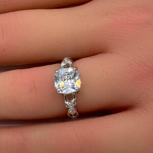 May include: A silver-toned ring featuring a large, clear, cushion-cut center stone. The band is crafted with a delicate, intricate metal design that extends from the base of the central setting, showcasing a polished finish and reflective facets.