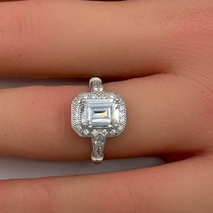 May include: A silver-toned ring featuring a central emerald-cut clear stone surrounded by a halo of smaller round stones. The band is slender and includes additional small stones set along the shoulders, creating a detailed, reflective appearance.