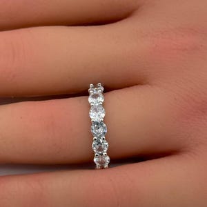 May include: A silver-toned band ring featuring five round, clear, faceted stones set in a row. The stones are held in place by small metal prongs, creating a continuous line of sparkle across the center of the band, which is worn on a finger.