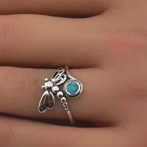 May include: A silver ring with a dragonfly motif and a turquoise gemstone. The ring is displayed on a hand with manicured nails. The dragonfly has detailed wings and a small, round turquoise stone.