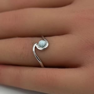 May include: A silver ring featuring a small, round, pale blue gemstone. The ring has a distinctive, swirling design. The ring is on a person's hand, with a white and pink background.