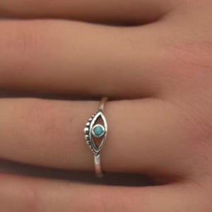 May include: A silver ring featuring a turquoise evil eye design. The ring is worn on a finger.