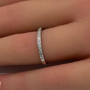 May include: A thin, silver-toned band ring featuring a row of small, clear, round-cut stones embedded along the top surface. The metal has a polished, reflective finish, and the stones are set closely together in a straight, uniform line.