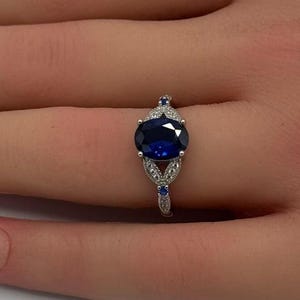 May include: A silver-toned ring featuring a large, oval-cut deep blue center stone. The band is accented with small, clear pavé-set stones and two tiny blue stones positioned at the top and bottom of the central setting, creating a floral-like design.