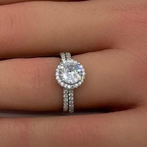 May include: A silver-toned ring featuring a large, round, clear center stone surrounded by a halo of smaller, sparkling stones. The band is thin and also encrusted with a row of tiny, clear stones, creating a continuous, brilliant, and reflective appearance.