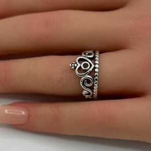 May include: A silver-colored ring with a crown design featuring heart and swirl details. The ring is worn on a finger, with a soft pink flower in the background.