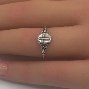 以下が含まれることがあります： 小さな丸い十字架のデザインが施された銀の指輪。指につけられています。