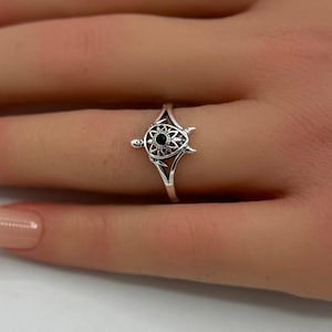 Op de afbeelding: Zilveren ring met een gedetailleerd schildpadontwerp en een zwarte middensteen. De ring wordt gedragen aan een hand met verzorgde nagels, tegen een zachte achtergrond met roze bloemen.