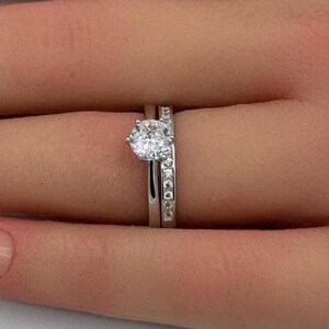May include: A silver-toned engagement ring set featuring a central round-cut clear stone held in a prong setting, paired with a matching band encrusted with a row of small, round, clear stones along the top half of the metal band.